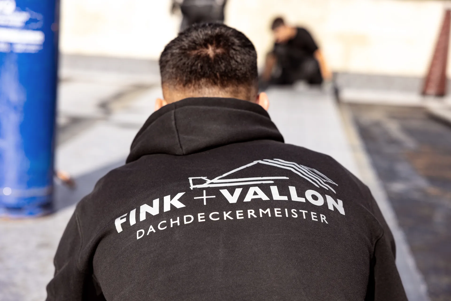 Der Rücken eines Mitarbeiters mit der Aufschrift „Fink + Vallon Dachdeckermeister“ auf der Arbeitsjacke ist zu sehen.