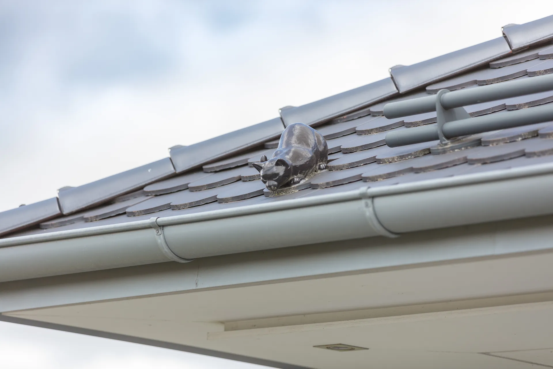 Auf einem Dach liegt eine graue Dachrinne, daneben sitzt als Schmuck eine Katze aus Metall.