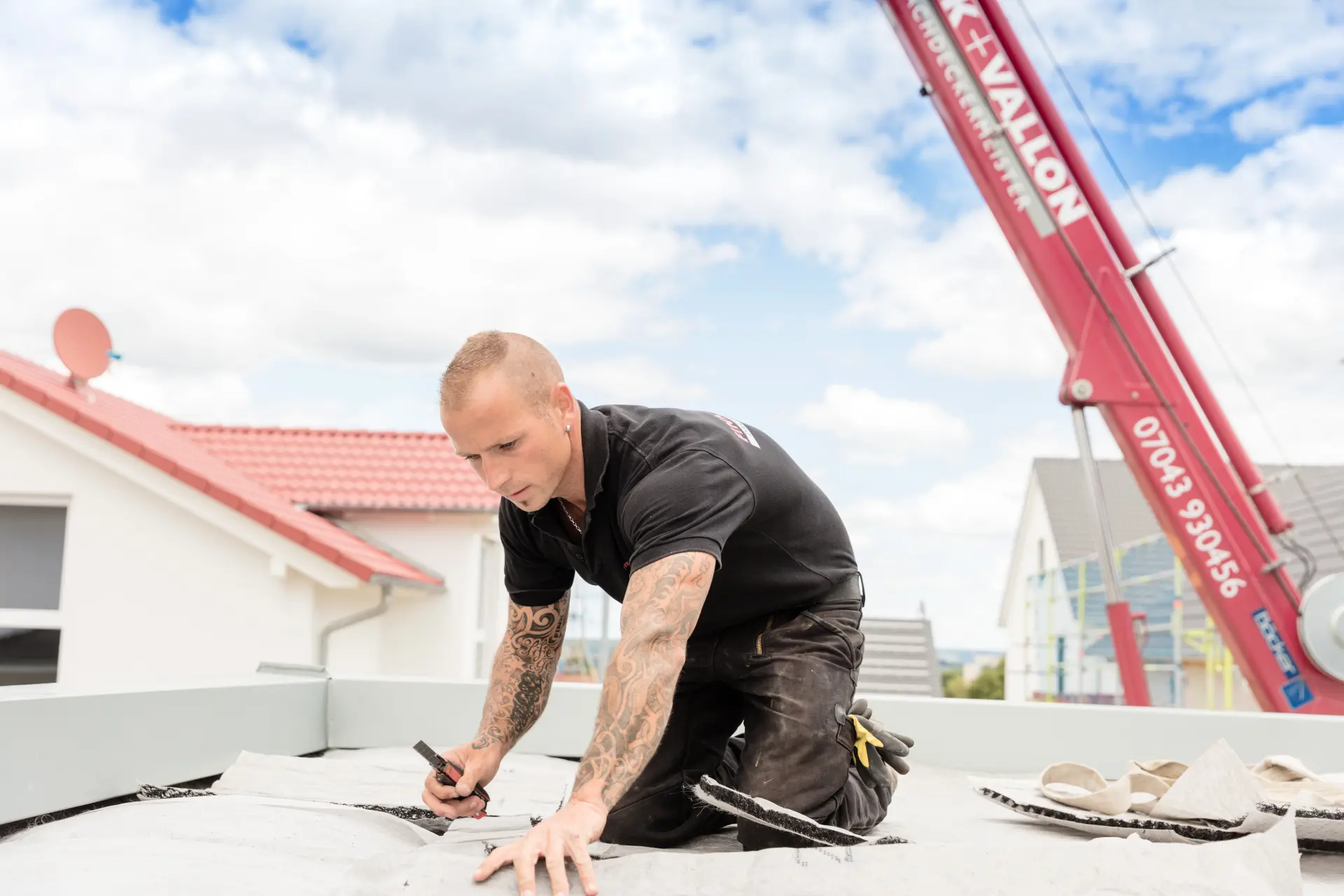 Ein Mann arbeitet kniend an einer Flachdachabdichtung, im Hintergrund ist ein roter Baukran.